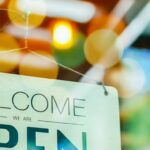 A hanging sign on a glass door reads "Welcome We Are Open," with colorful lights and reflections behind it.