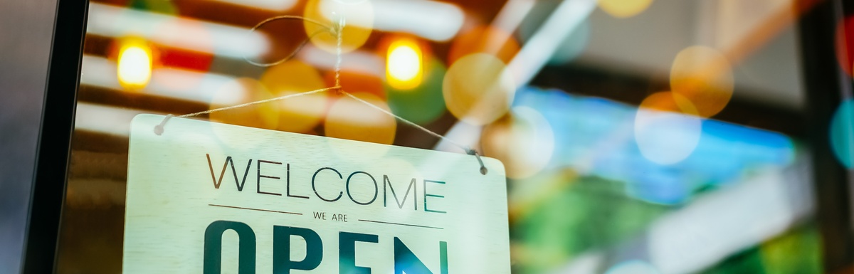 A hanging sign on a glass door reads "Welcome We Are Open," with colorful lights and reflections behind it.