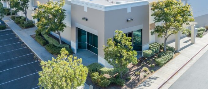 The sun shines on a large, one-story industrial office building with landscaped beds and an empty parking lot.