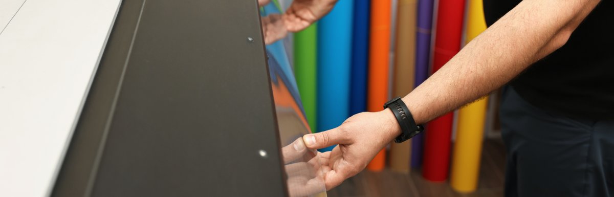 A man using a wide-format printer opens a panel on the machine. There are large rolls of paper ranging in several colors.
