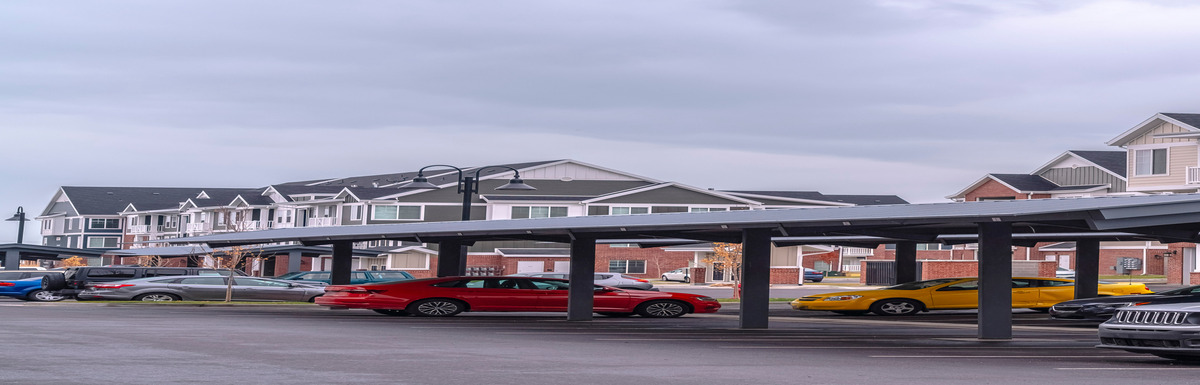 Several cars with different paint colors parked underneath a metal carport with apartment buildings in the background.