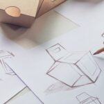 A person using a pencil to sketch the dimensions and different perspectives of a cardboard container with a handle.