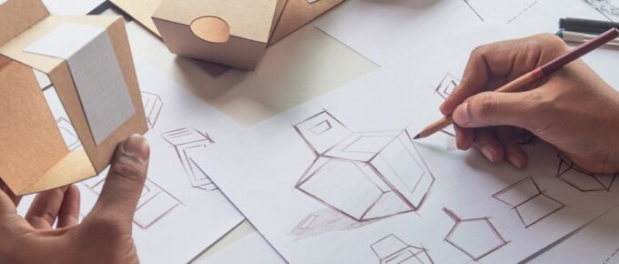 A person using a pencil to sketch the dimensions and different perspectives of a cardboard container with a handle.