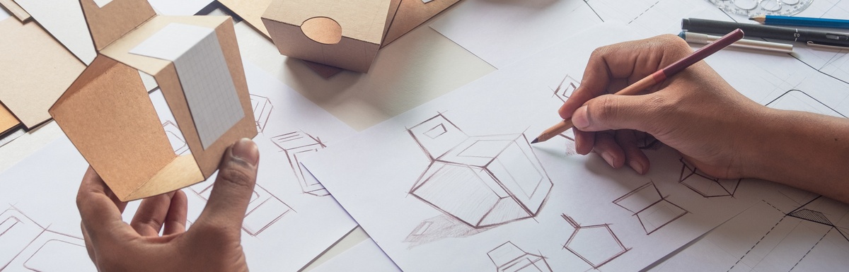 A person using a pencil to sketch the dimensions and different perspectives of a cardboard container with a handle.