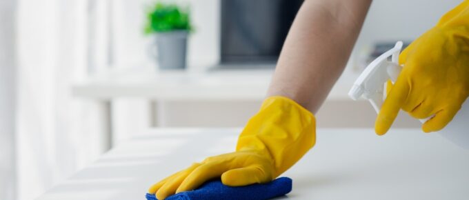 Hands in yellow gloves hold a spray bottle and a blue cloth as they clean a white table inside an office environment.