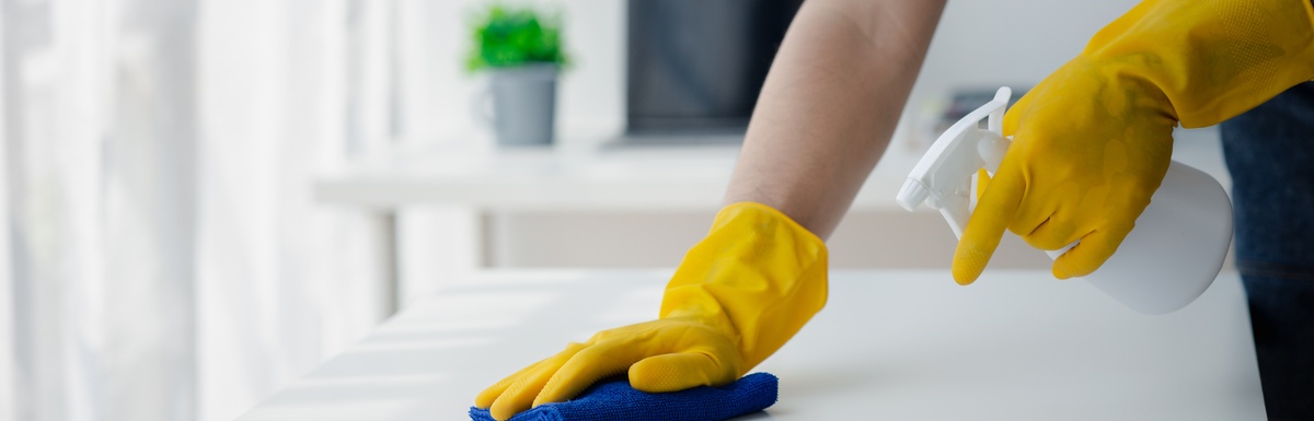 Hands in yellow gloves hold a spray bottle and a blue cloth as they clean a white table inside an office environment.