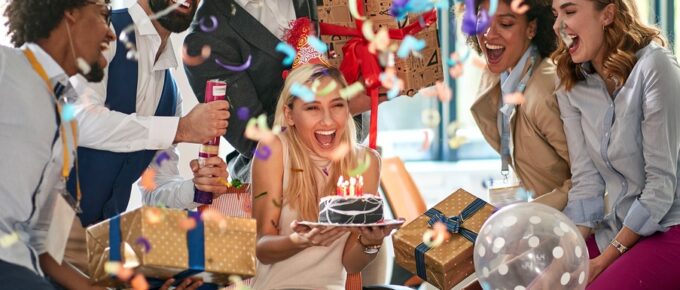 Coworkers wearing party hats and holding presents surround an employee sitting in an office chair holding a small cake.