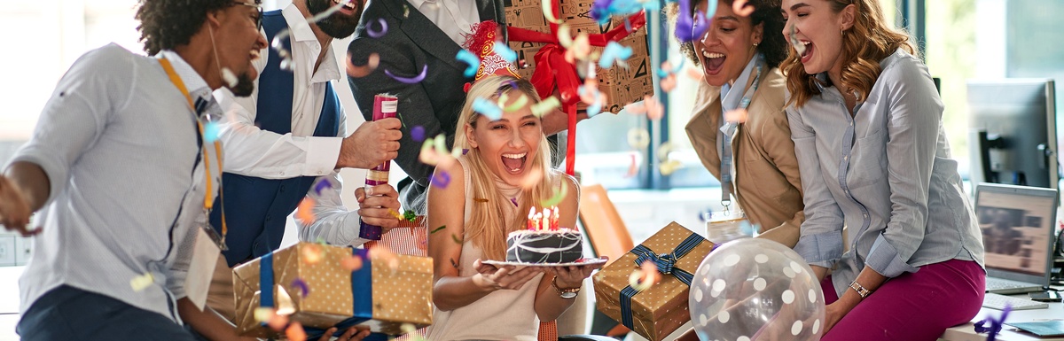 Coworkers wearing party hats and holding presents surround an employee sitting in an office chair holding a small cake.