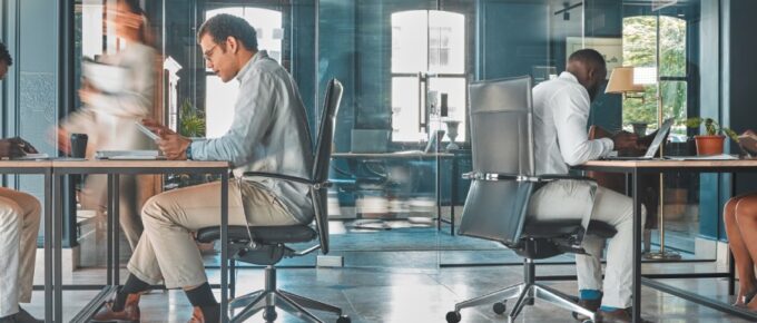Men and women inside an office with a glass wall and a large window. One walks while the others work at desks.