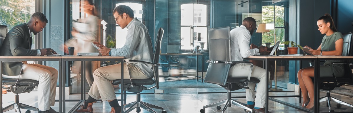 Men and women inside an office with a glass wall and a large window. One walks while the others work at desks.