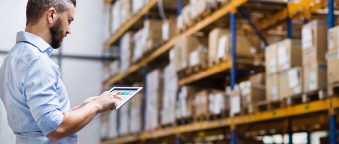 A middle-aged man in a collared shirt standing inside a large warehouse full of boxes. He's reading a tablet.