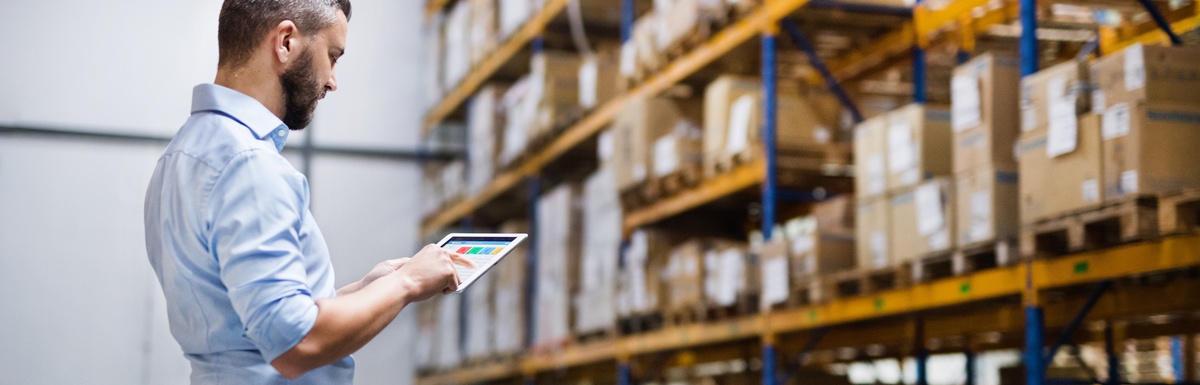 A middle-aged man in a collared shirt standing inside a large warehouse full of boxes. He's reading a tablet.