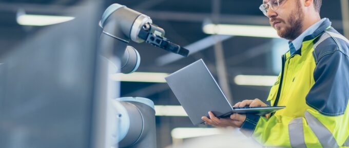 A man wearing a yellow safety jacket holds an open laptop while standing in front of a robotic machine.