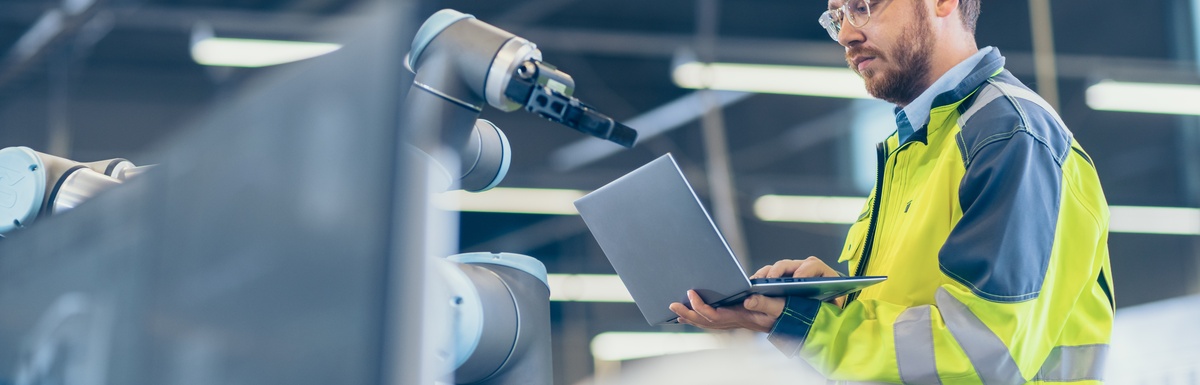 A man wearing a yellow safety jacket holds an open laptop while standing in front of a robotic machine.