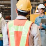 Three people in construction gear face two individuals dressed professionally, wearing hard hats, as they converse.