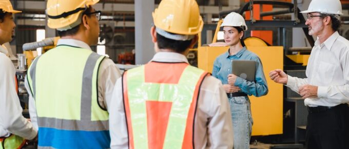 Three people in construction gear face two individuals dressed professionally, wearing hard hats, as they converse.