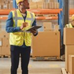 A man wearing a hard hat and a yellow reflective vest. He's looking up, holding a tablet and surrounded by carton boxes.