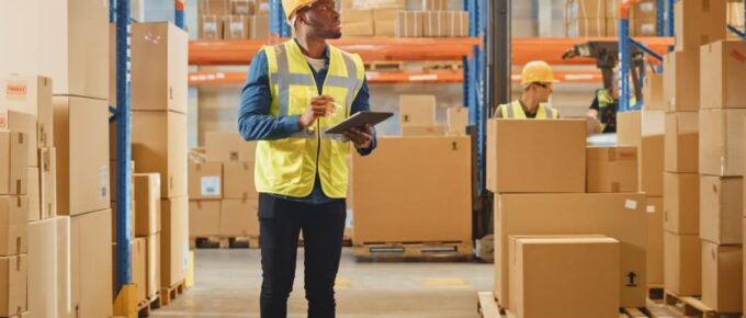 A man wearing a hard hat and a yellow reflective vest. He's looking up, holding a tablet and surrounded by carton boxes.