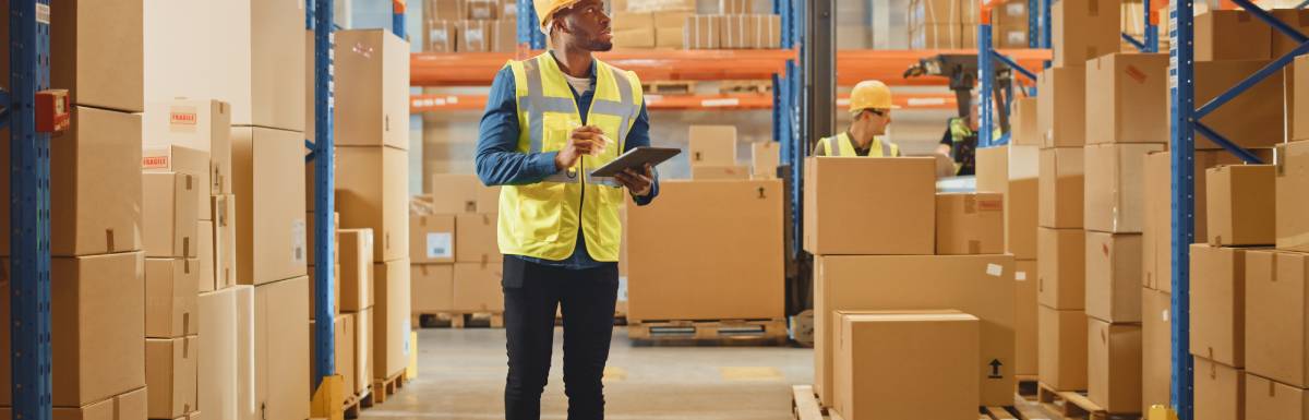 A man wearing a hard hat and a yellow reflective vest. He's looking up, holding a tablet and surrounded by carton boxes.