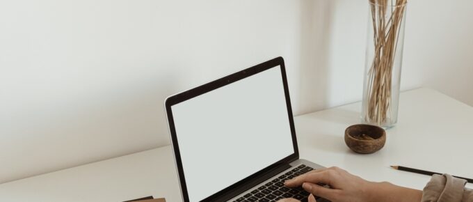 Two hands resting on a laptop keyboard that sits on a white desk next to a brown notebook and plant in a vase.