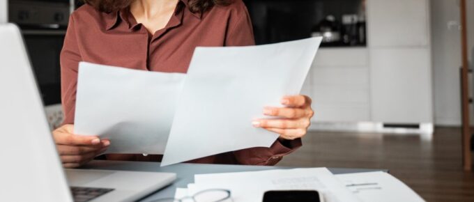 A woman in a brown shirt looks at two pieces of paper while sitting in front of a laptop on a desk with other papers.