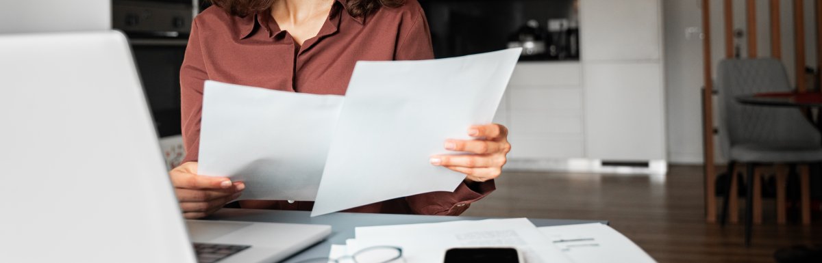 A woman in a brown shirt looks at two pieces of paper while sitting in front of a laptop on a desk with other papers.