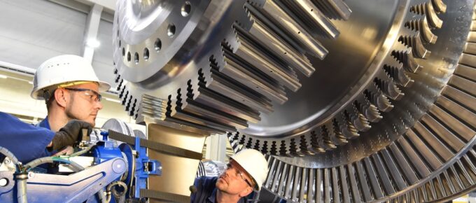 In an industrial setting, two workers assemble a steam turbine. Both workers are wearing blue overalls and hard helmets.