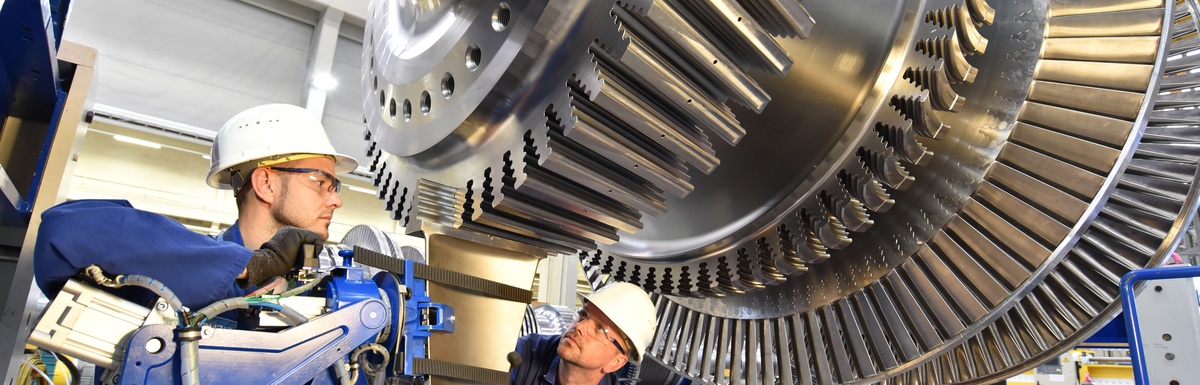 In an industrial setting, two workers assemble a steam turbine. Both workers are wearing blue overalls and hard helmets.