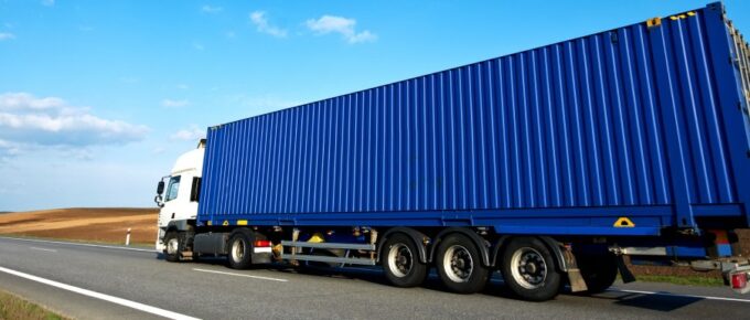 A semi rides down a road with a blue cloudy sky in the background. The trailer of the semi is blue.