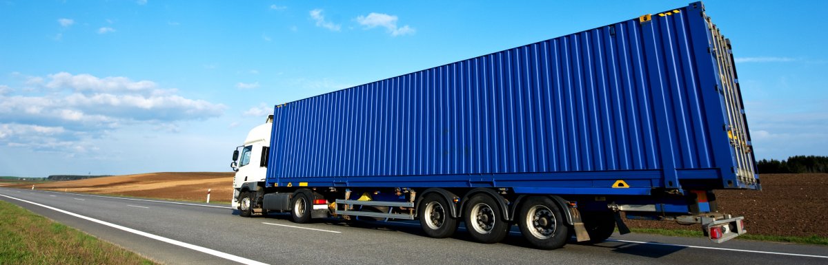 A semi rides down a road with a blue cloudy sky in the background. The trailer of the semi is blue.