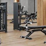 The inside of a modern gym with a black ceiling, wooden beams, and several exercise machines and benches on the floor.