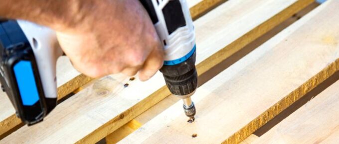 A man using a blue-and-white drill to insert a screw into the hole of a wooden pallets surface board.
