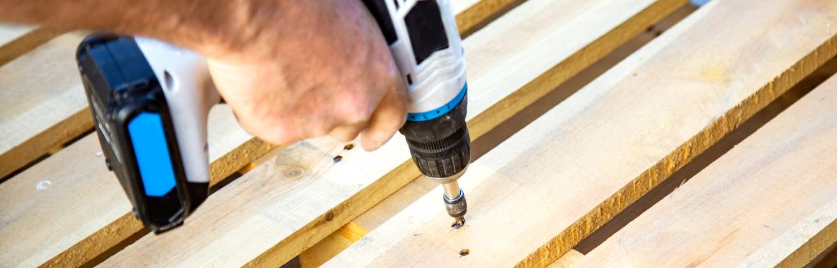 A man using a blue-and-white drill to insert a screw into the hole of a wooden pallets surface board.