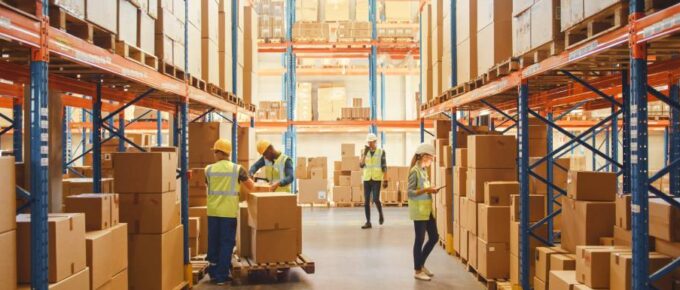 One long aisle of a retail warehouse is lined with boxes on high shelves. Employees inspect and catalog the boxes.