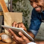 A businessman is smiling, standing in front of bread, as he uses a tablet device for online business orders at his bakehouse.