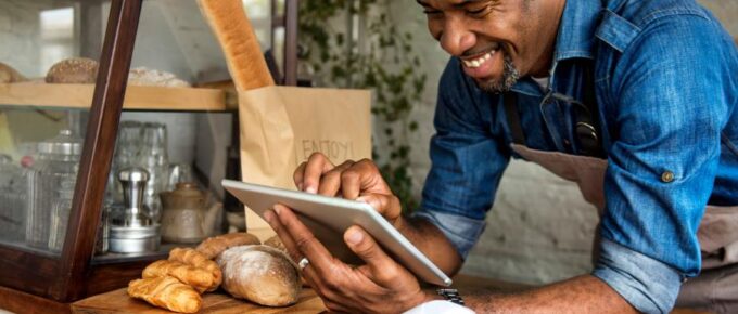 A businessman is smiling, standing in front of bread, as he uses a tablet device for online business orders at his bakehouse.