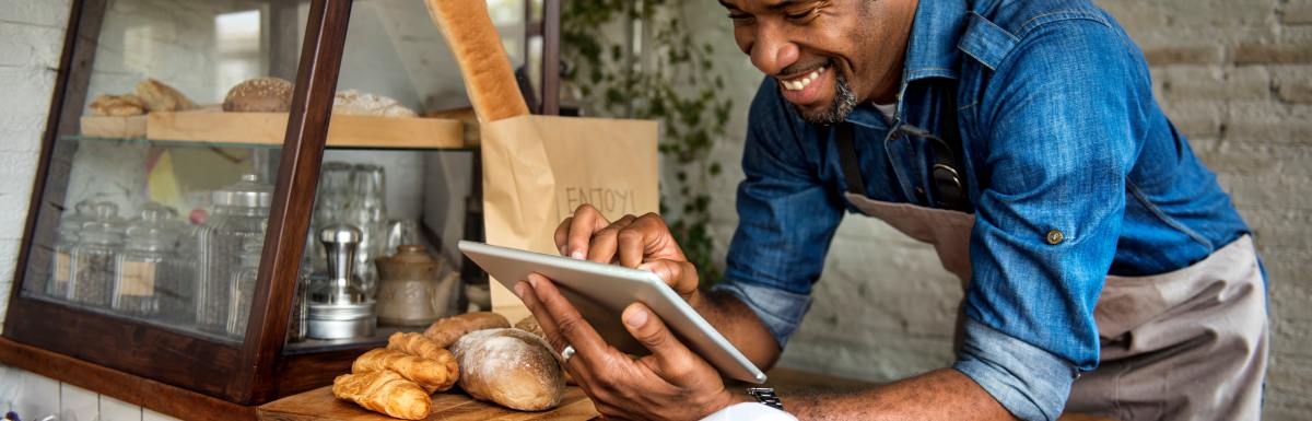 A businessman is smiling, standing in front of bread, as he uses a tablet device for online business orders at his bakehouse.