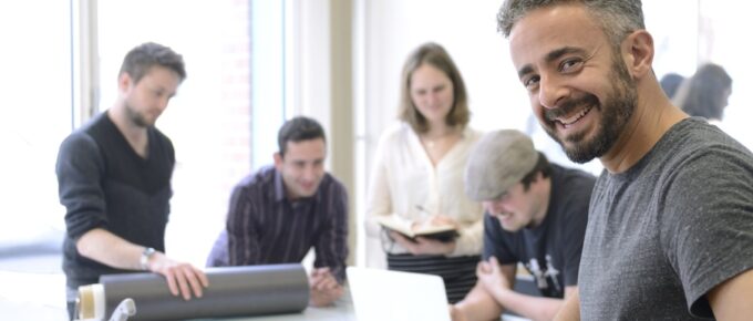 A business owner in a gray shirt smiles as his team members work in the background. They stand in front of glass walls.