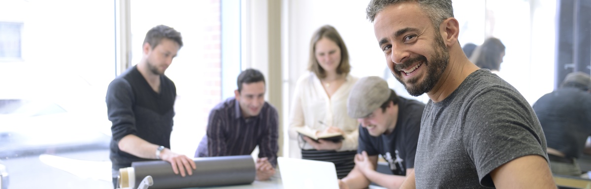 A business owner in a gray shirt smiles as his team members work in the background. They stand in front of glass walls.