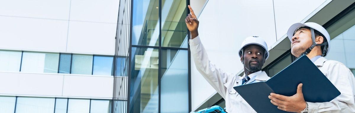 Two engineers with hard hats on, inspecting a building. One of them points at something, getting their coworker's attention.
