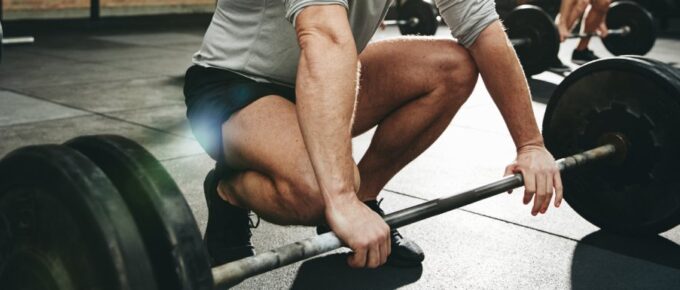 A man looks down and smiles as he squats to grab a barbell with plated weights on it. Behind him, another man does the same.