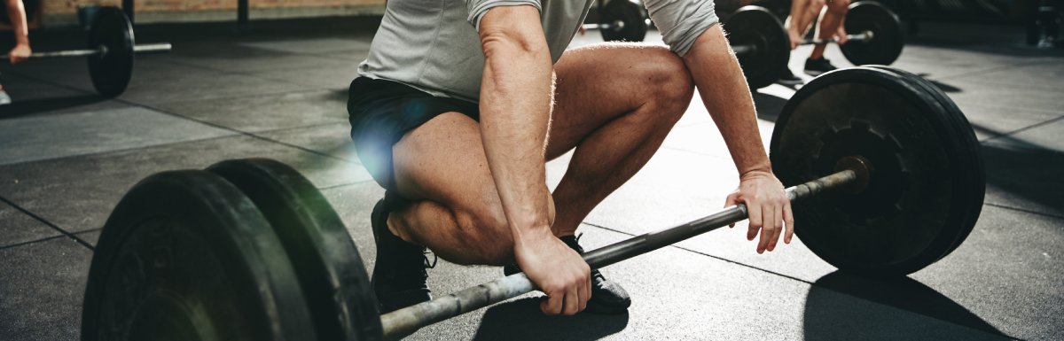 A man looks down and smiles as he squats to grab a barbell with plated weights on it. Behind him, another man does the same.