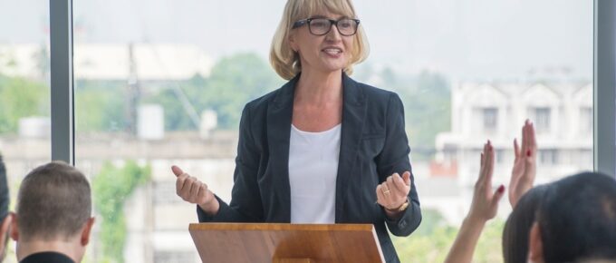 A woman stands at a wooden podium and speaks to a crowd of young professionals with large glass windows behind her.