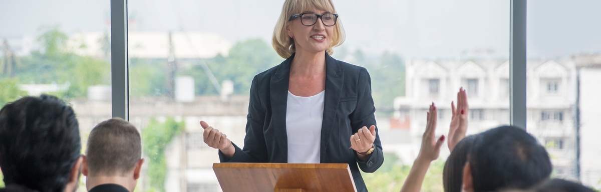 A woman stands at a wooden podium and speaks to a crowd of young professionals with large glass windows behind her.