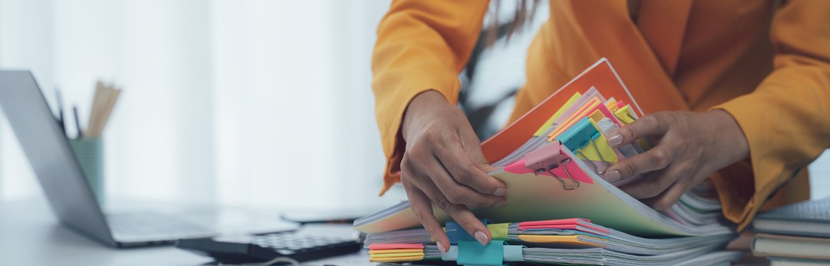 A person wearing a suit jacket sifts through stacks of paper separated with color-coded binder clips and sticky notes.