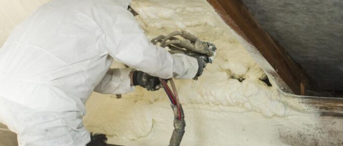 A person wearing a white protective suit kneels and applies polyurethane spray foam on the roof of an attic.