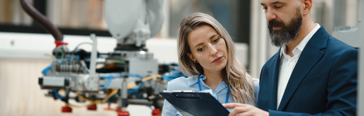 A man in a business suit showing a woman a clipboard. They are standing in a manufacturing facility.