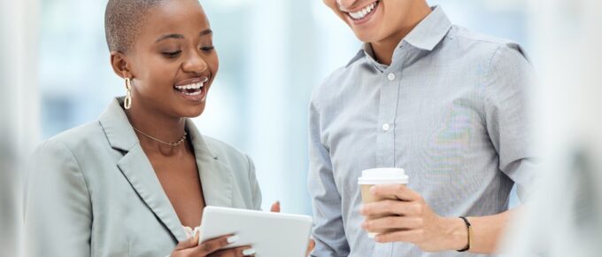 A woman holds a tablet and stands beside a man in a button-down shirt. Both smile and look at the tablet screen.
