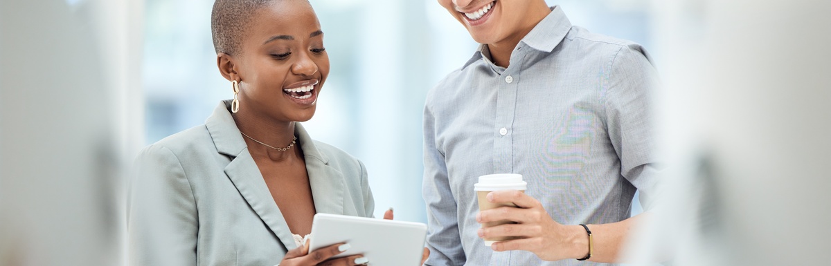 A woman holds a tablet and stands beside a man in a button-down shirt. Both smile and look at the tablet screen.