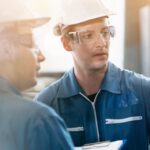 Two industrial workers in hard hats review machinery, one pointing at a control panel while the other holds a clipboard.